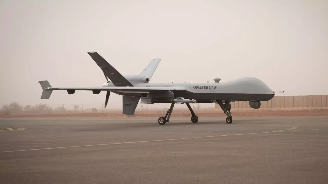 O dronă Reaper gri a Forțelor Aeriene Franceze, la sol, pe o pistă gri, cu un gard în plan secundar.