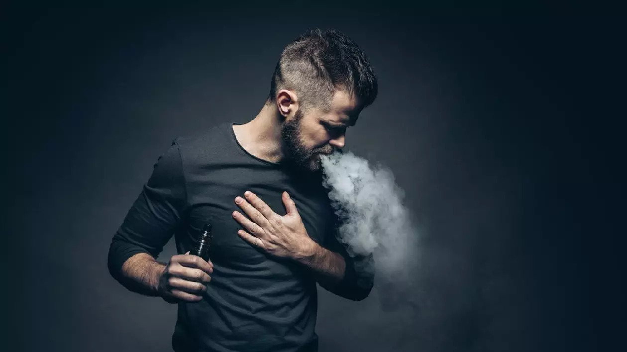 Un tânăr îmbrăcat în bluză neagră, pe fundal negru, vapează dintr-o tigară electronică- Vaporizarea e-lichidului aromatizat de la o țigară electronică