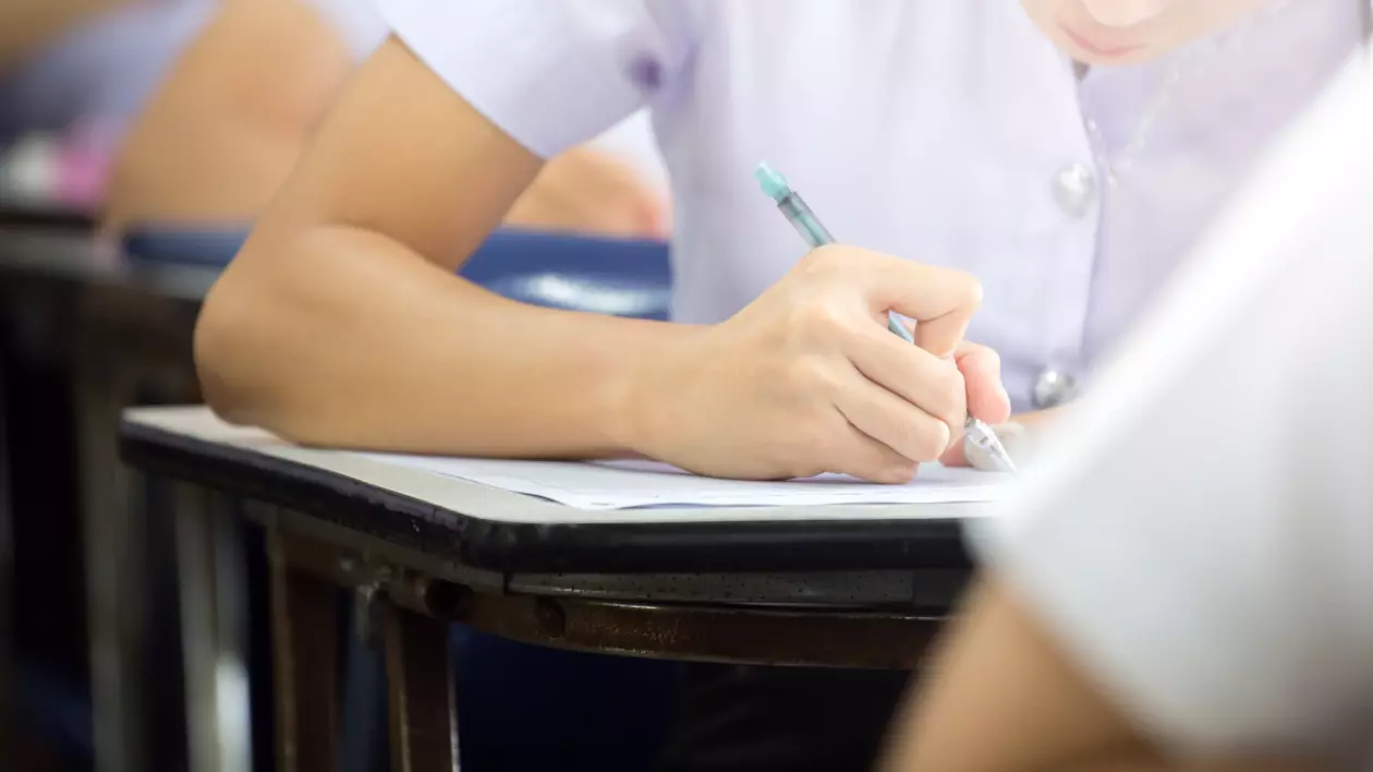 Mai muți elevi, surpinși de la gât în jos, stau în bănci unul în spatele celuilalt, și scriu pe foi de hârtie, în timpul unui examen.