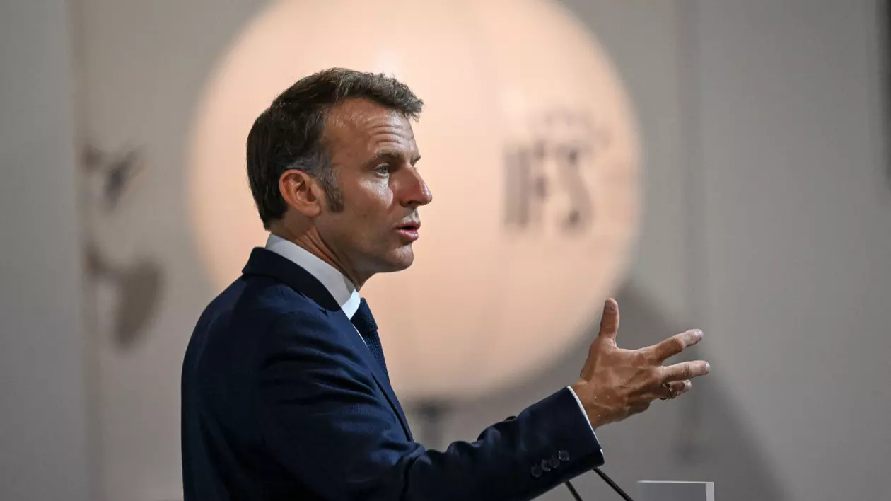 În imagine este ilustrat Emmanuel Macron, în costum negru, în timp ce ține un discurs