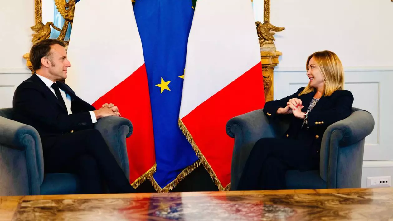 Întâlnire între prim-ministrul italian Giorgia Meloni și președintele Franței, Emmanuel Macron. Stau față în față, pe scaune și zâmbesc. În spatele lor, steagul Italiei și steagul Uniunii Europene.