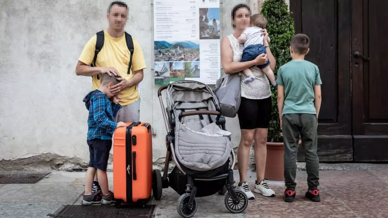 Un român cu fața blurată îi acoperă ochii băiețelului care ține mâna pe un geamantan portocaliu, lângă ei este un cărucior de copii, soția cu fața blurată ține un bebeluș în brațe, iar lângă ea se află băiatul cel mare, fotografiat cu spatele. Este o familie de români din Italia care a rămas fără casă
