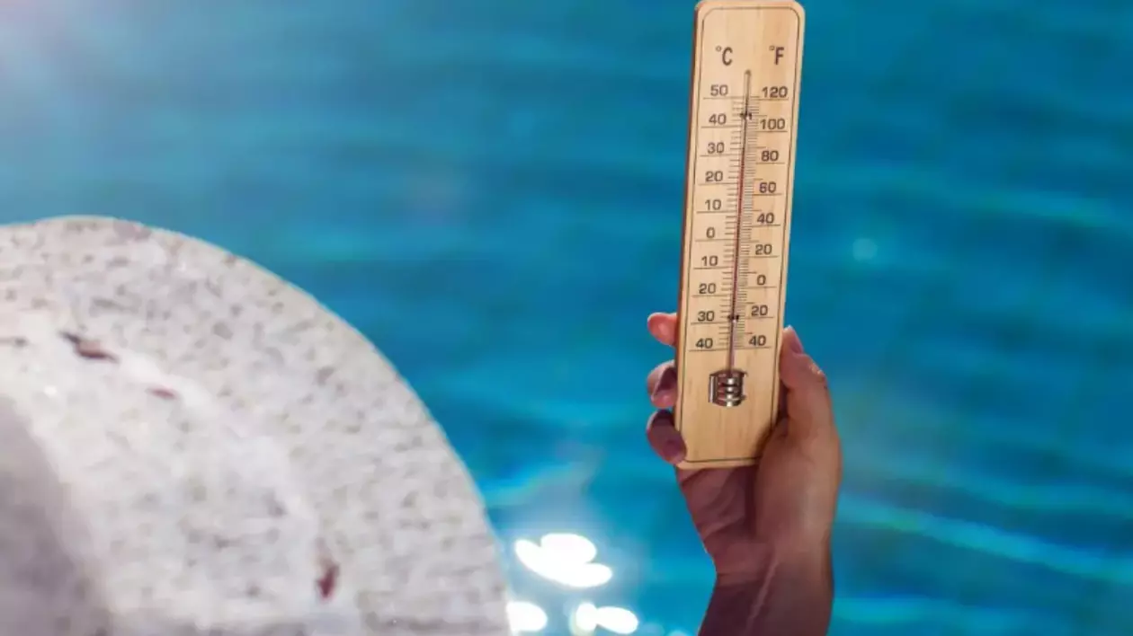 În imagine este ilustrată o femeie care stă la piscină, are o pălărie albă pe cap și ține un termometru în mână