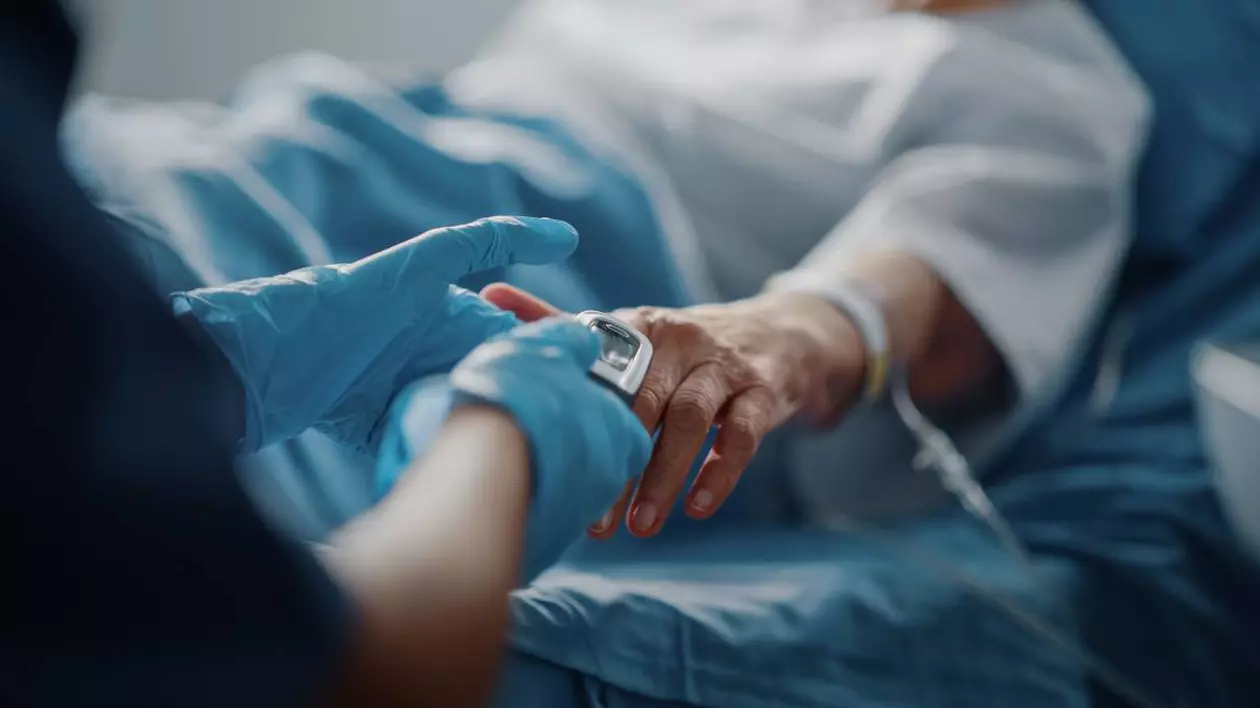 O femeie în vârstă pe patul de spital, după o intervenție, are puls oximetru montat la mână, iar o asistentă o îngrijește.