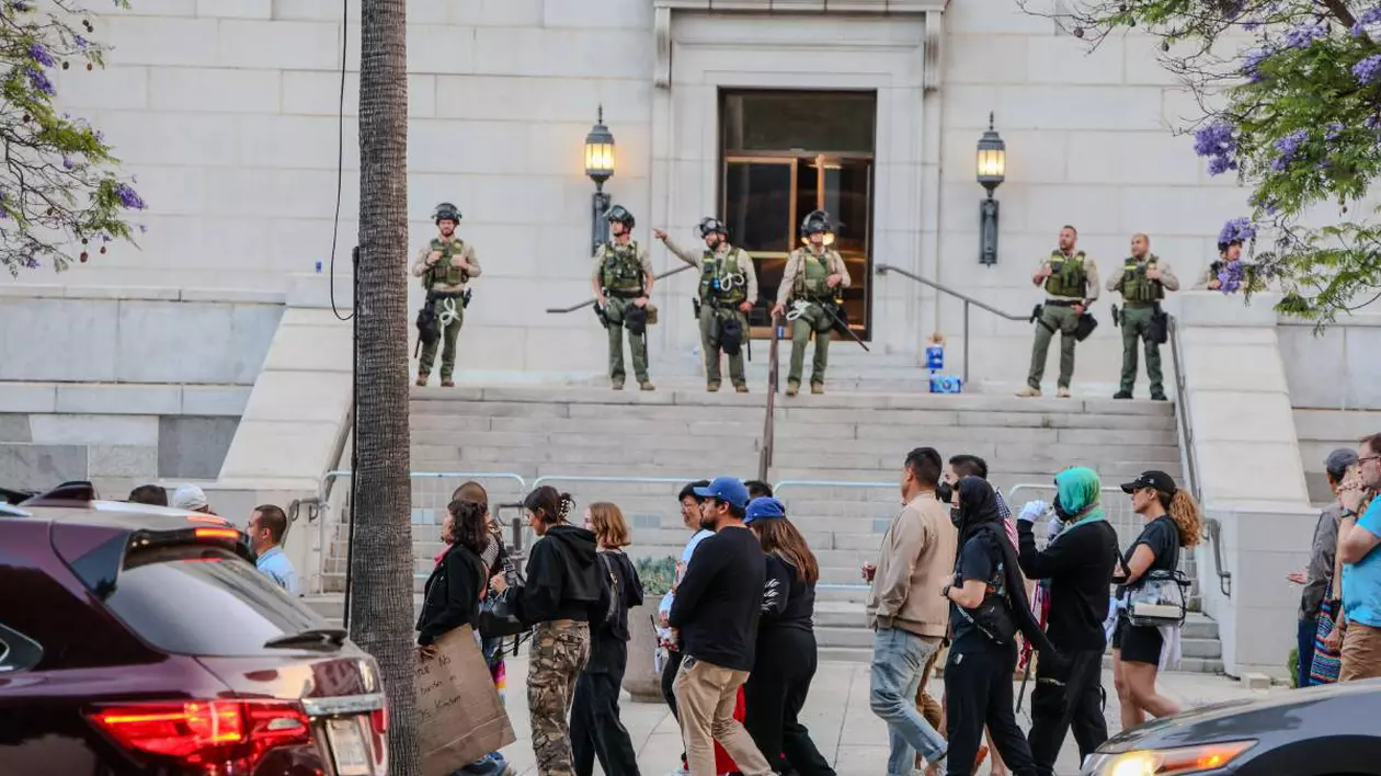 Militari, pe treptele unei clădiri din Los Angeles, în timpul protestelor, în fața lor se află un grup de tineri protestatari care stau pe trotuar.