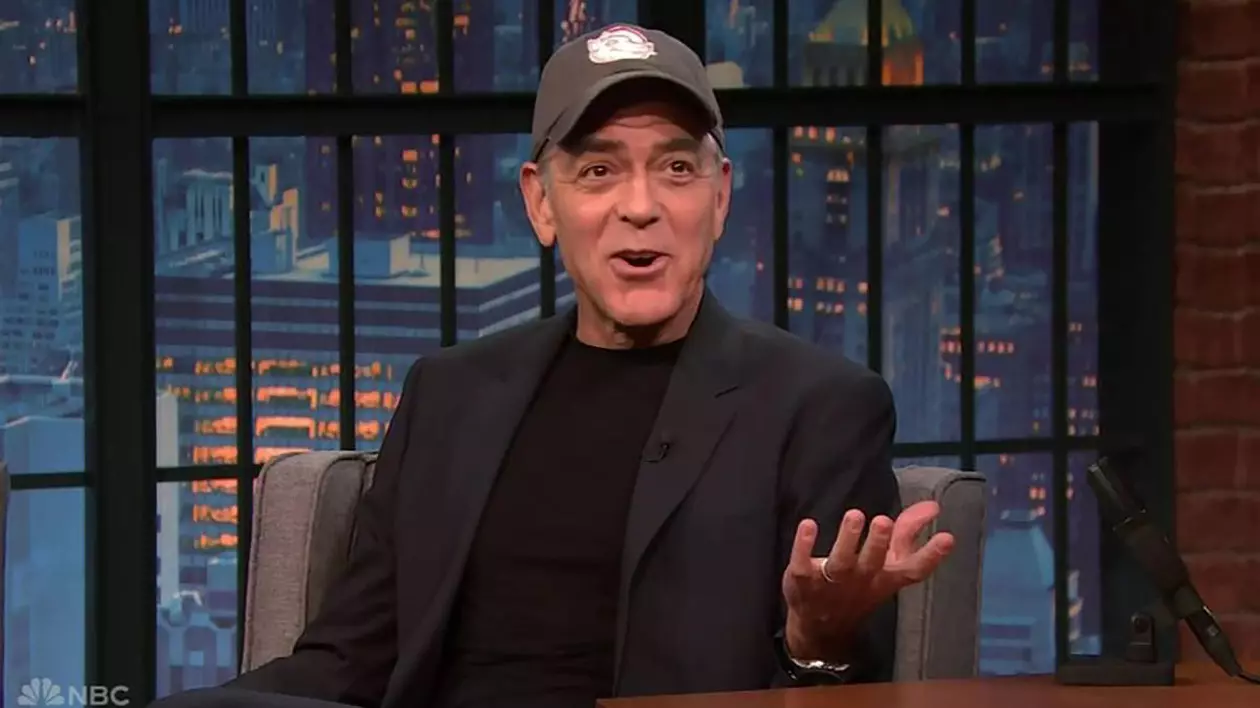 George Clooney în timpul unei emisiuni TV, stă pe canapea într-un costum negru, gesticulează cu mâinile și poartă o șapcă pe cap.