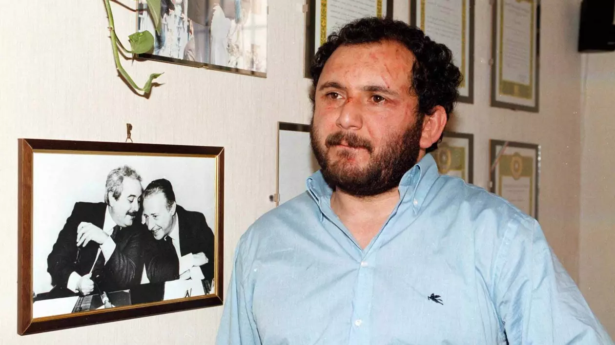 Giovanni Brusca poartă o cămașă albastră, iar în spatele său sunt atârnate pe perete fotografii