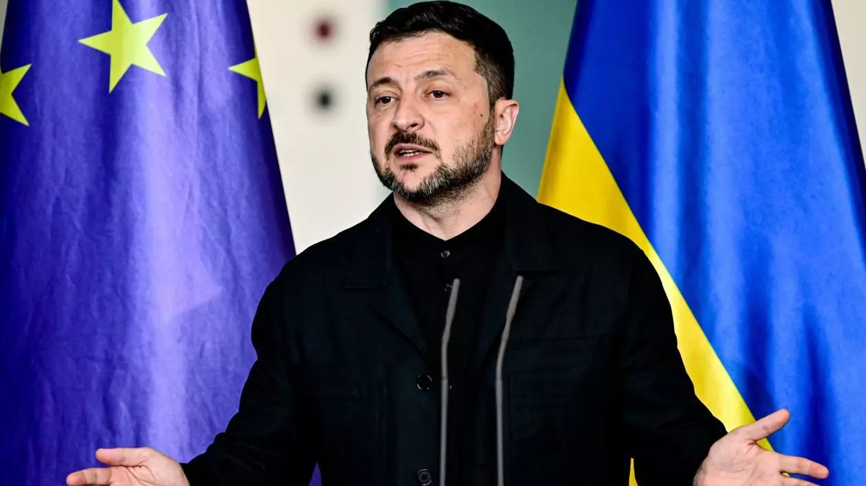 Volodimir Zelenski, îmbrăcat într-o cămașă neagră, dă explicații cu mâinile larg deschise în timpul unei conferințe de presă, având în spate steagurile Ucrainei și al Uniunii Europene