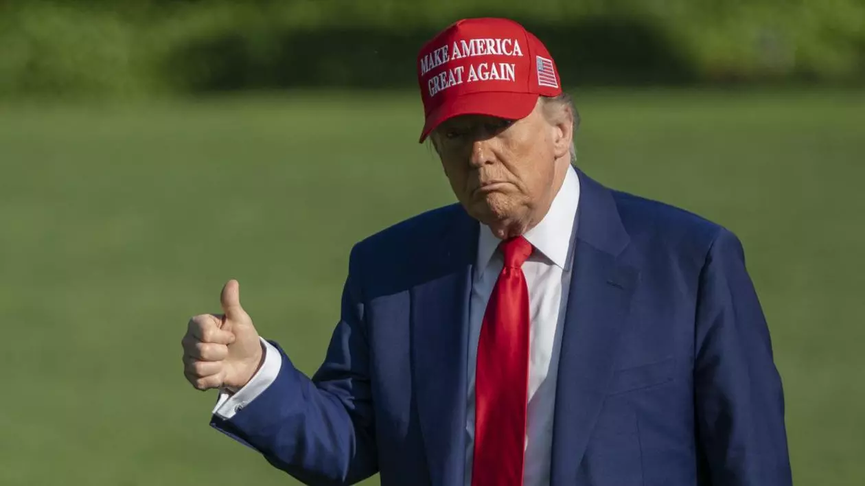 Donald Trump, îmbrăcat la costul gri, cu cravată rși și purtând pe cap o șapcă roșie cu mesajul „Make America Great Again”, ține degetul mare ridicat pe peluza din fața Casei Albe
