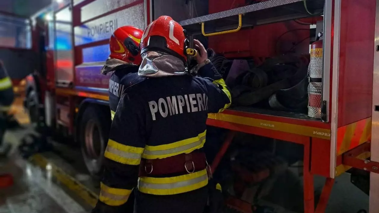 Doi pompieri echipați și cu căști roșii pe cap în prim-plan, fotografiați cu spatele, în timp ce umblă în autospeciala de stingere
