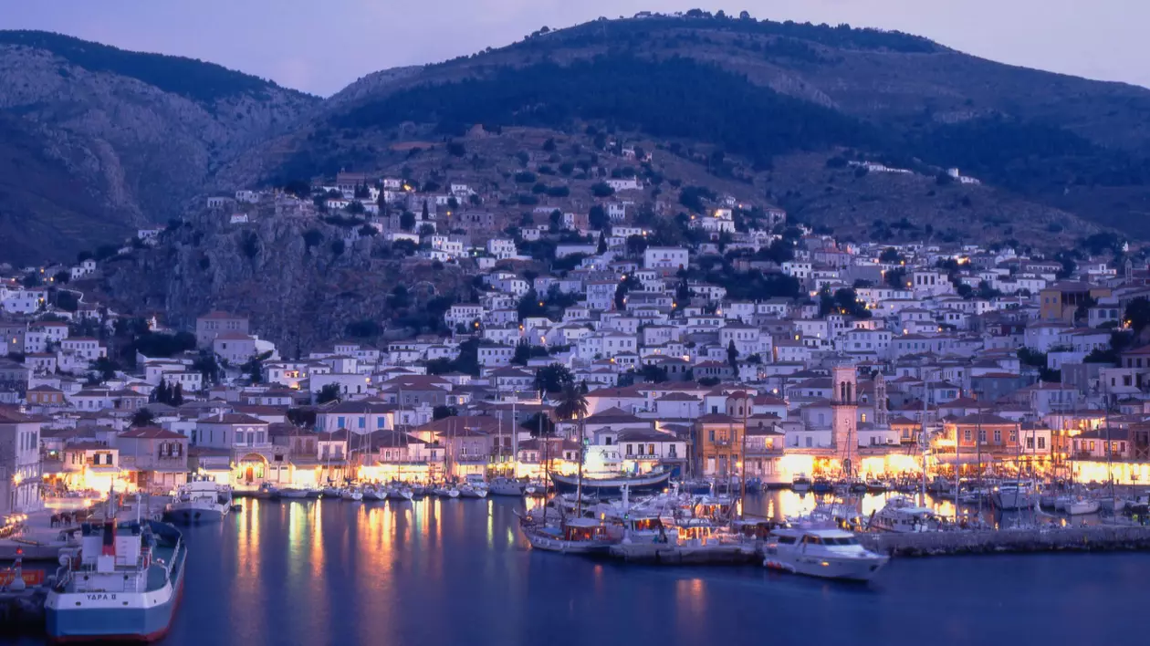 Fotografia surprinde portul insulei Hydra, Grecia, la lăsarea serii, cu luminile clădirilor reflectându-se în apa liniștită.