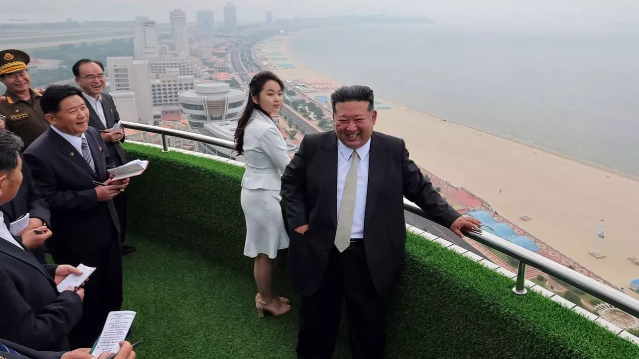 Fotografie publicată de Agenția Centrală de Știri Coreeană îl prezintă pe liderul nord-coreean.Kim Jong-un, în costum negru si camasă albă, și fiica sa, Kim Ju Ae, intr-un deux-piece alb, vizitează zona turistică de coastă Wonsan Kalma, in stanga lor numerosi jurnalisti, in spate se vede plaja si mare intr-o zi cu ceață. 24 iunie 2025