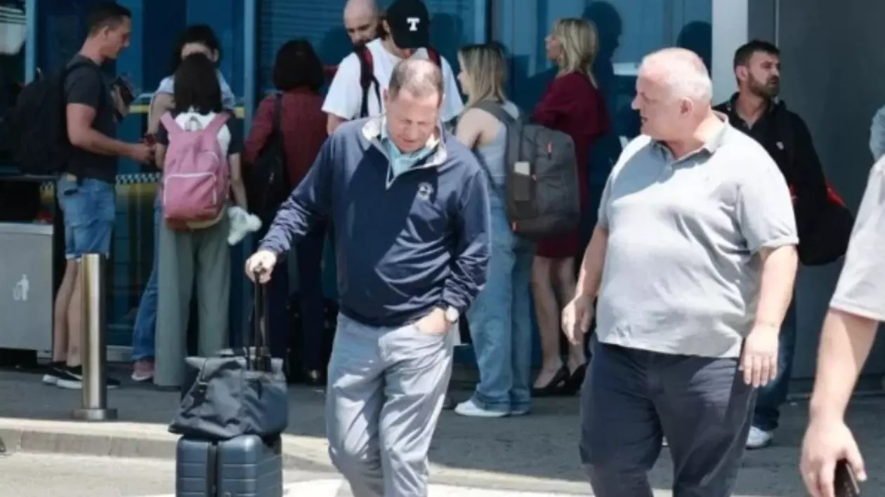 În prim-plan se află doi bărbați: unul cu un troller și o geantă de mână, îmbrăcat într-o jachetă sport și pantaloni deschiși la culoare, care a aterizat recent; celălalt poartă un tricou gri și pantaloni închiși la culoare, și îl privește pe primul.