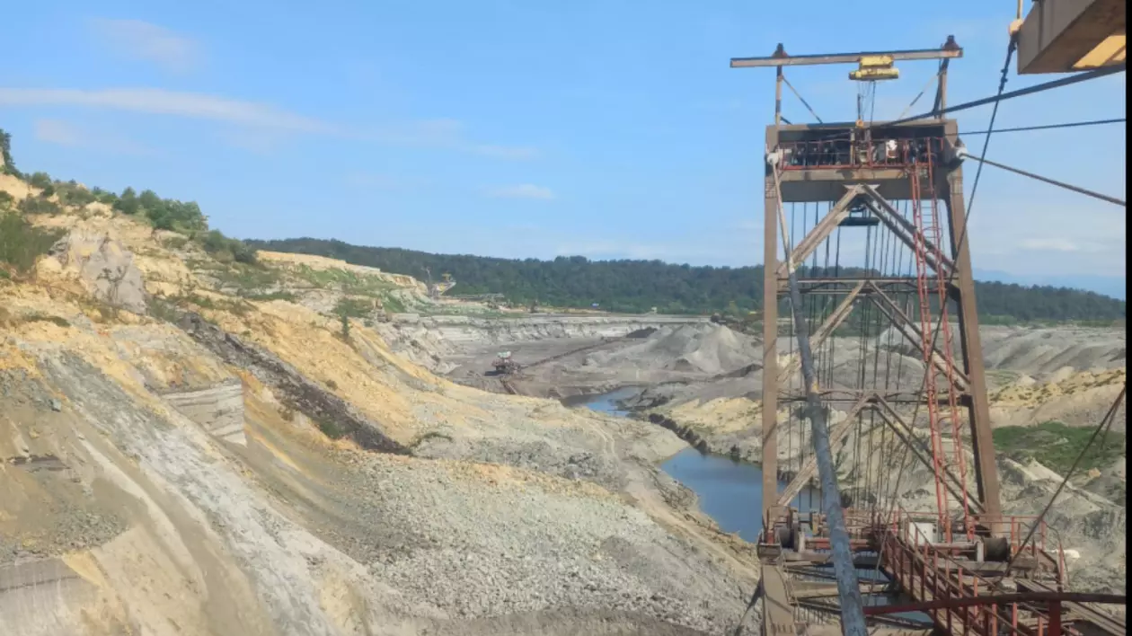 Imaginea prezintă un peisaj industrial, unde o linie de transport a cărbunelui traversează un teren excavat.