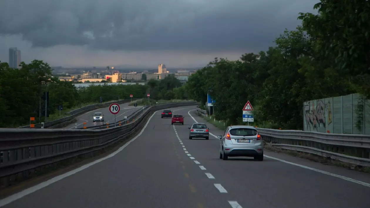 Mai multe mașini circulă pe un drum cu două benzi, cerul este înnorat