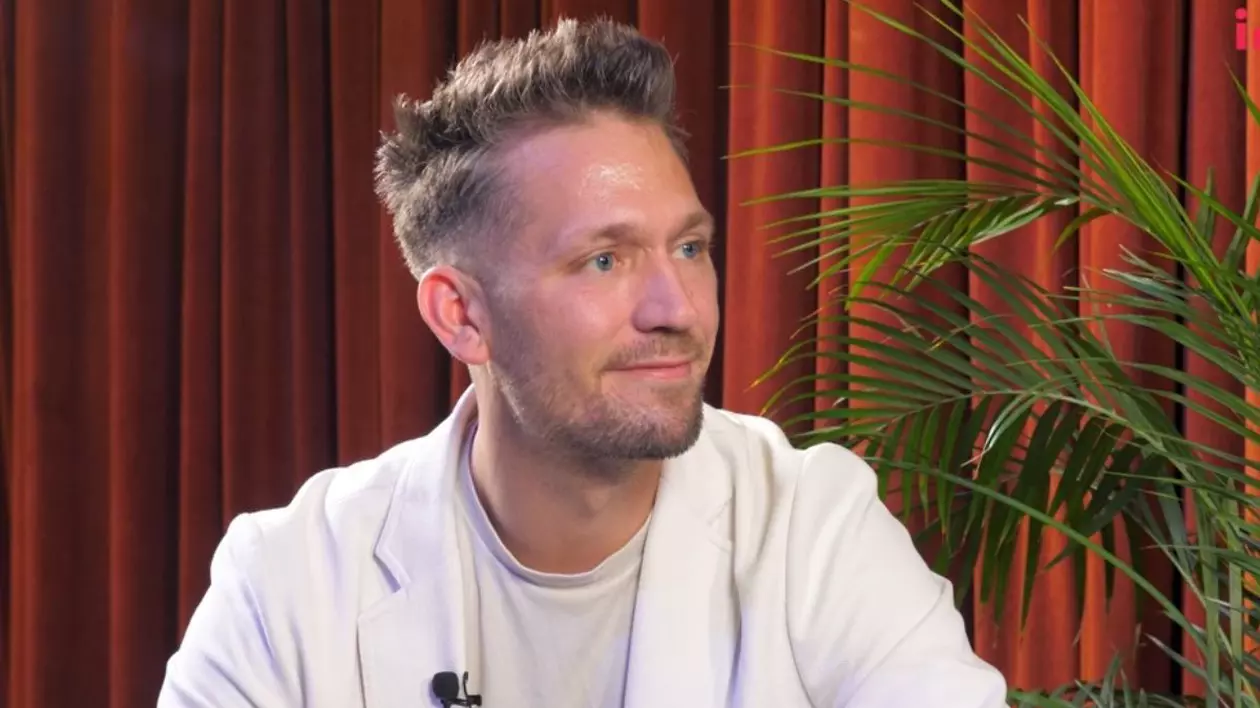 Michael Topolinski, îmbrăcat cu un tricou alb și un sacou alb, în timp ce stă pe fotoliu, în platoul Interviurile Imobiliare.ro, cu o perdea roșie pe fundal și o plantă verde