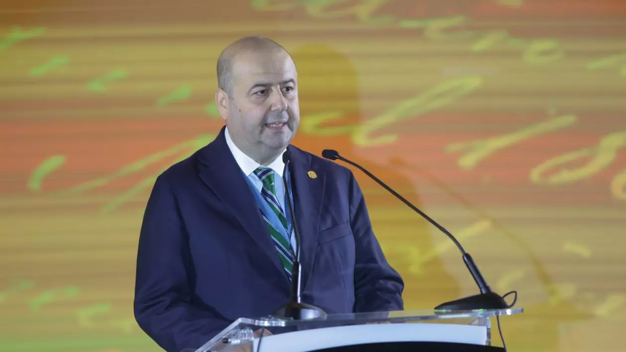 Mihai Busuioc, in costum inchis la culoare si cravata in dungi nuante de albastru si bleu, in timp ce tine un discurs la un eveniment aniversar, in spate un ecran mare in nuante de galben si portocaliu, septembrie 2024