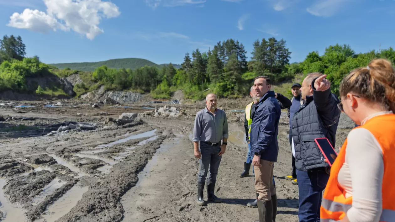 Imaginea surprinde o vizită de teren într-o zonă afectată de inundații. Grupul de persoane – format din oficiali și specialiști – se află pe un teren noroios, cu urme adânci de utilaje și bălți de apă.