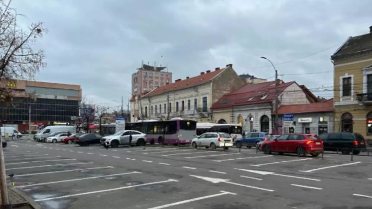 Imaginea surprinde parcarea din Piața Mihai Viteazul din Cluj-Napoca, într-o zi mohorâtă. Se pot observa mai multe mașini parcate, autobuze în stație.