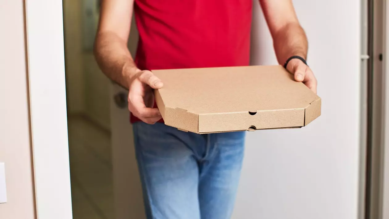 Imagine cu un bărbat, care poartă un tricou roșu și blugi, ținând în mâini o cutie cu pizza