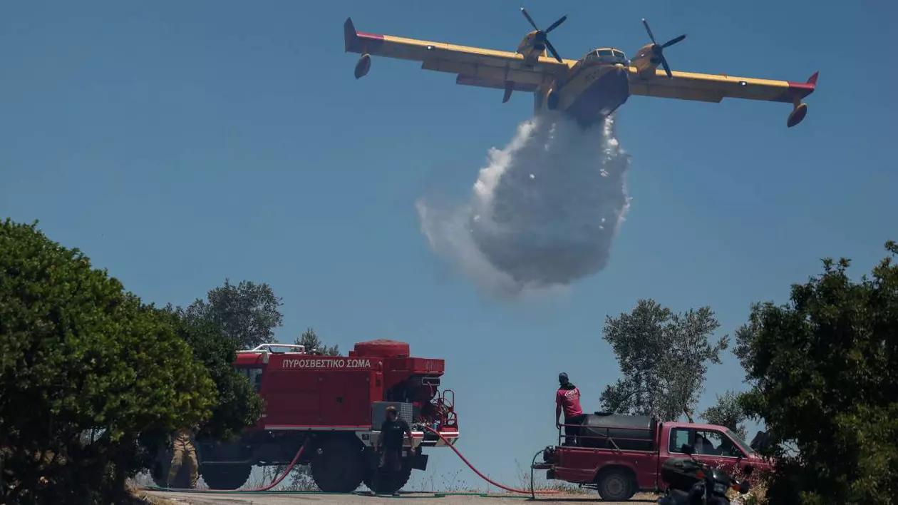 Un avion de stingere survolează o pădure din Grecia și aruncă apa peste vegetația în flăcări în timp ce echipajele de pompieri stau lângă pădure și așteaptă să intervină.