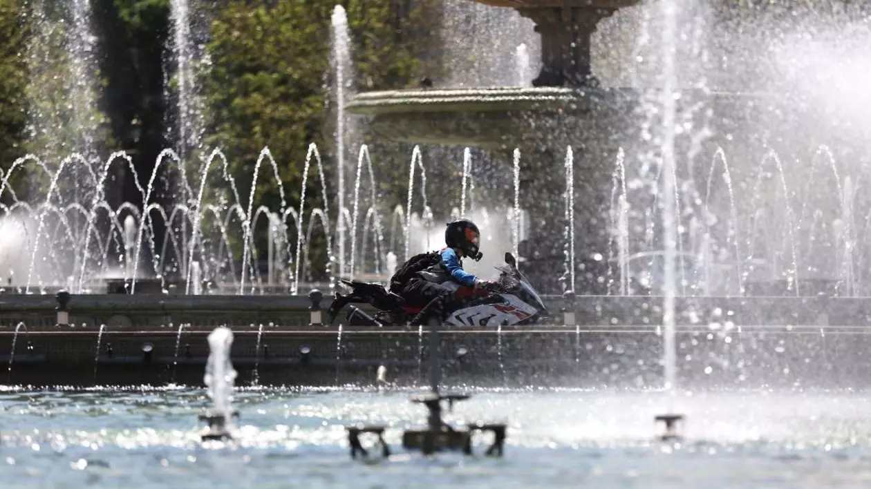 O fântână arteziană și șuvoaie de apă curgând, prin față trece un motociclist, iar în spate se văd niște copaci verzi, București, 24 aprilie 2025