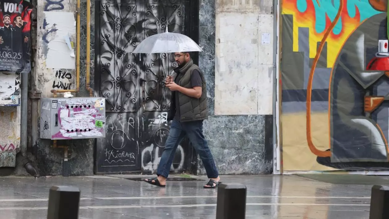 Un bărbat cu tricou cu mânecă scurtă și încălțat cu șlapi ține în mână o umbrelă deschisă deasupra capului și se apără de ploaie, mergând pe trotuar, în București, 26 mai 2025
