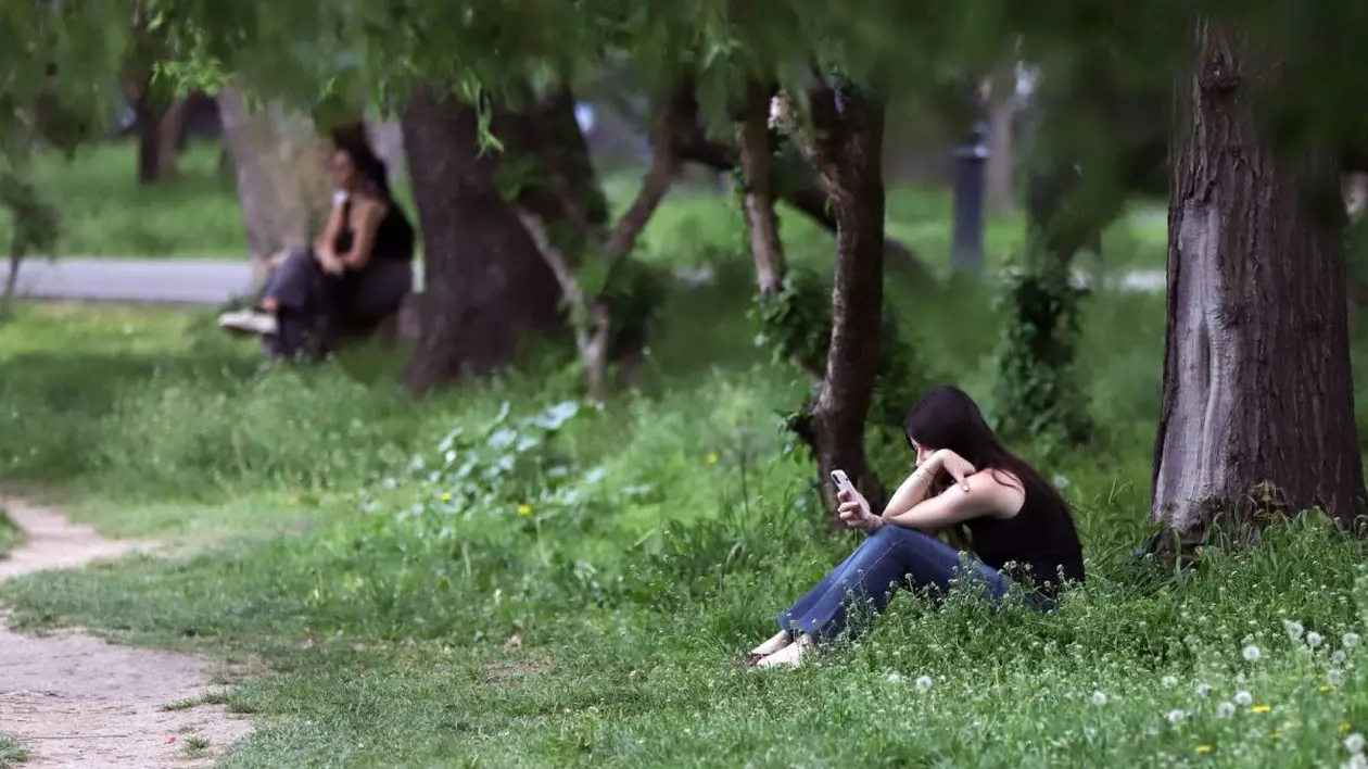 O fată cu maiou negru stă pe iarbă într-un parc și citește pe telefon, iar lângă ea se văd în spate doi tineri pe o bancă, București, parcul Cișmigiu, 25 aprilie 2025