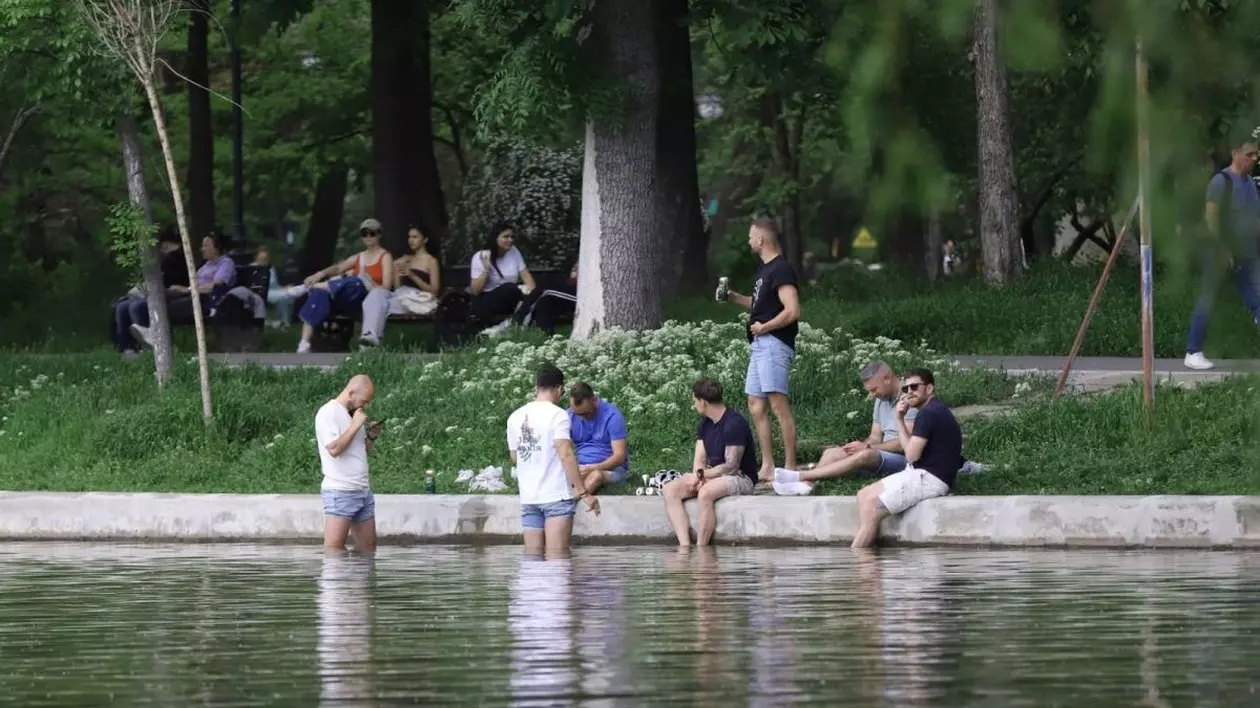 Doi băieți stau în picioare în apă, alți trei stau pe bordura lacului și cu picioarele în apă, un altul stă în picioare pe iarbă cu o doză în mână, iar în spate se mai văd câteva tinere și alți oameni care sunt pe bancă, în parcul Cișmigiu, București, 25 aprilie 2025