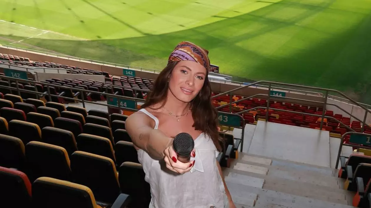 Raluka, îmbrăcată cu un costum lejer alb și o bandana colorată pe Arena Națională, ține microfonul în mână, în timpul repetițiilor pentru momentul în care va cânta imnul României.