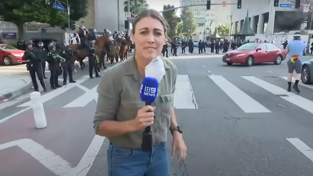 femeie blonda, in blug si camasa, cu un microfon in mana, in mijlocul unor proteste, forte de ordine in spatele ei,