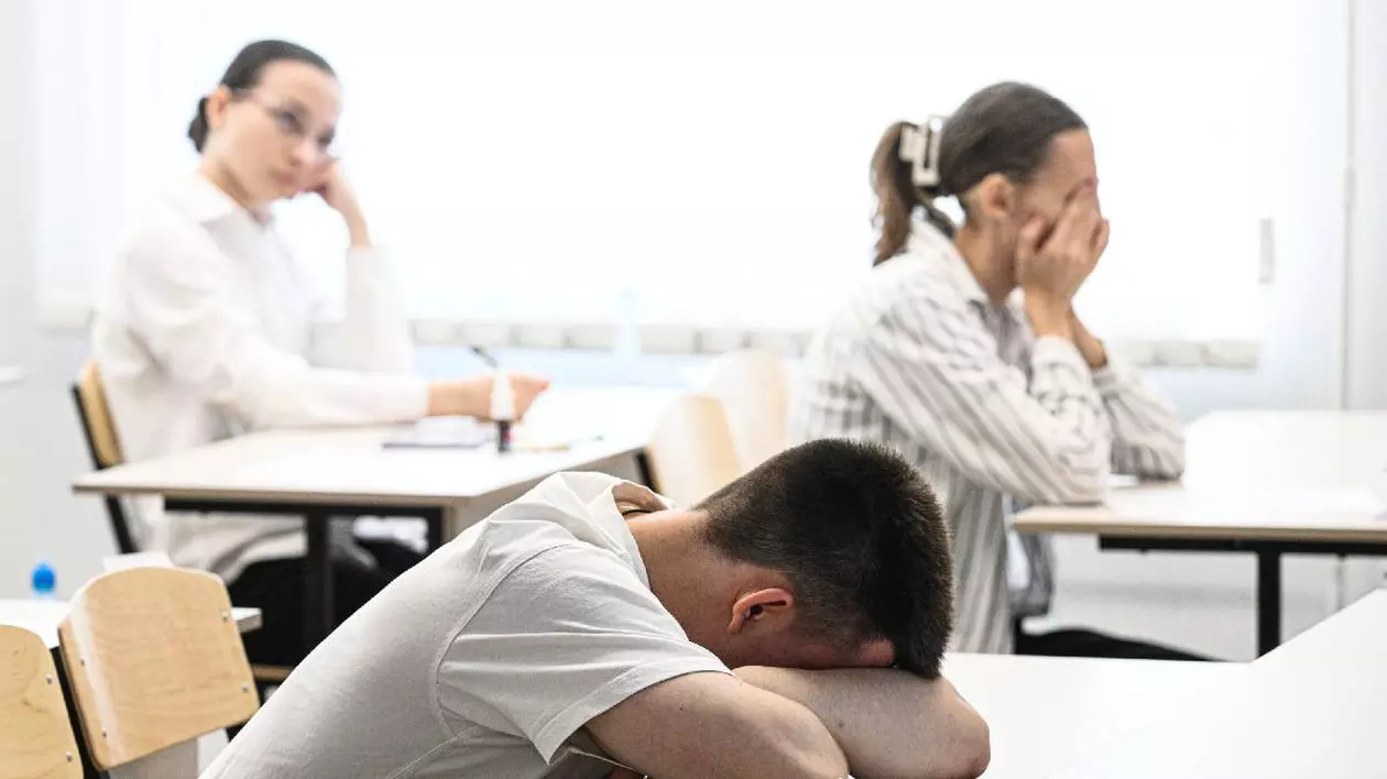 În imagine un elev stă cu capul sprijinit de mâini, pe bancă, o elevă își ține mâinile pe față dezamăgită iar o altă elevă privește spre partea din față a sălii de examen așteptând