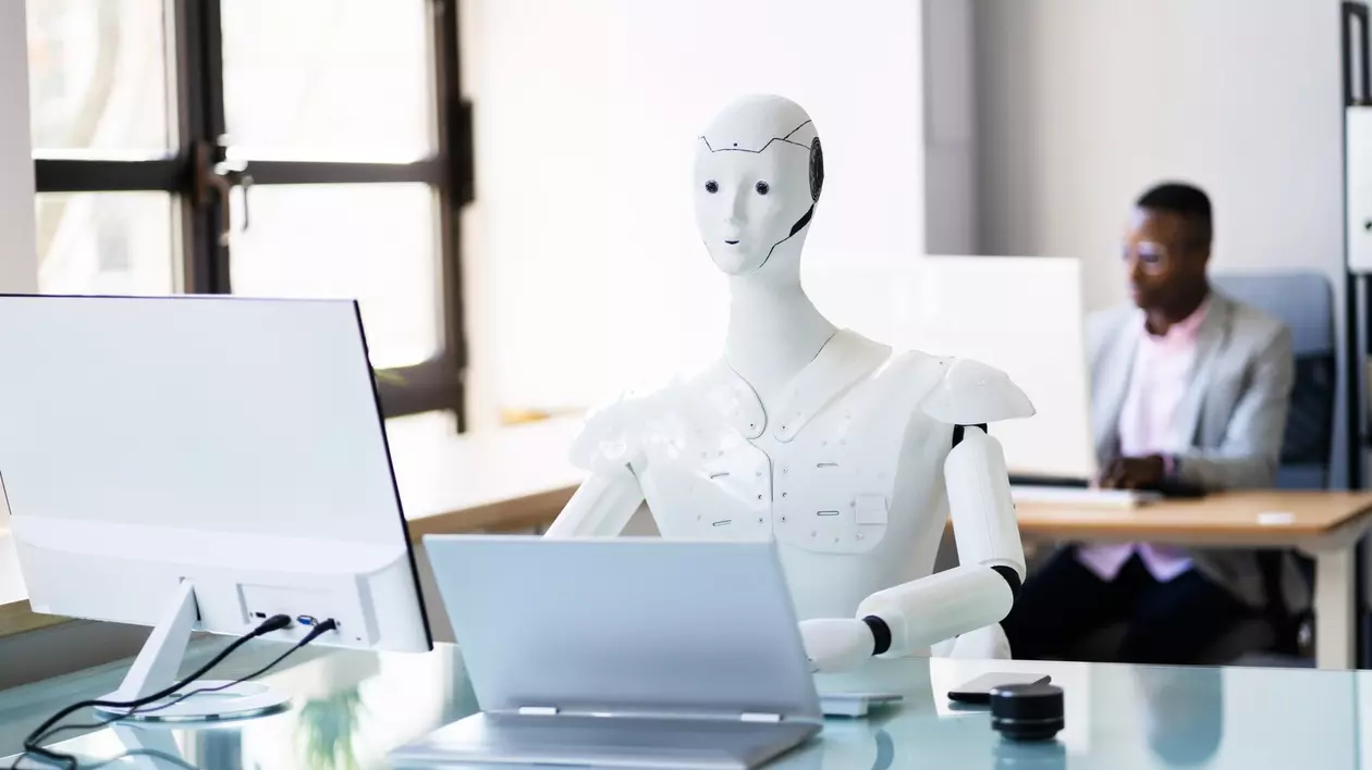 Imagine cu un robot alb stând la birou în fața unui laptop și a unui monitor. În spate se află un alt om care privește într-un monitor