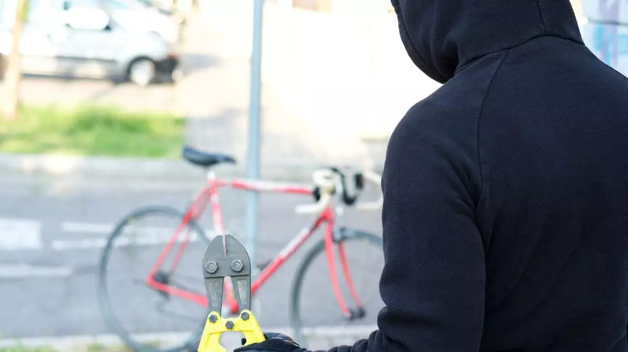 Un bărbat îmbrăcat cu un hanorac negru și cu gluga pe cap are un patent galben în mâna stângă și se uită la o bicicletă roșie parcată aproape de el