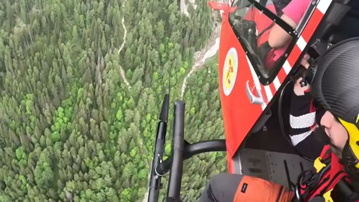 Imagini din elicopter cu salvamontist imbracat in rosu care se pregateste de coborare pe troliu pentru o operațiune de salvare deasupra unei paduri verzi