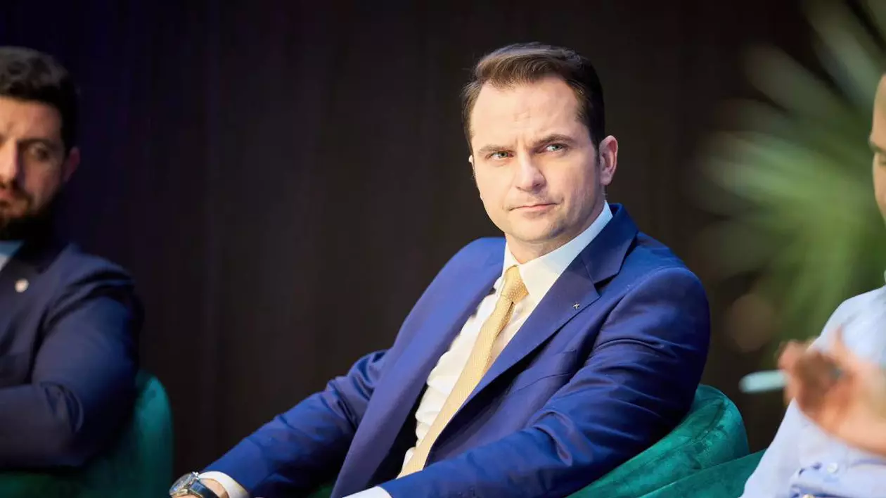 Ministrul Energiei, Sebastian Burduja, stând pe scaun în timpul unei conferințe, poartă un costum albastru și o cravată galbenă