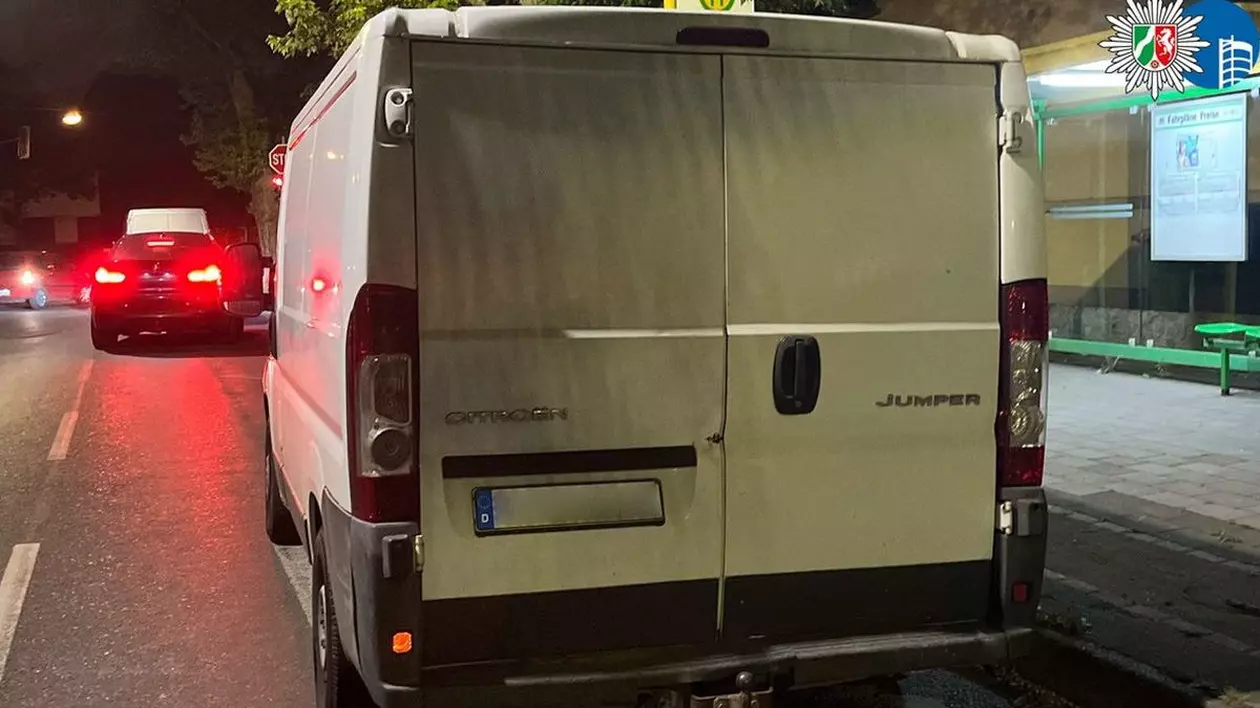 O mașină dubiță albă, fotografiată cu spatele și numărul de înmatriculare blurat, parcată pe o stradă din Germania, iar în fața ei se vede o mașină neagră cu farurile aprinse