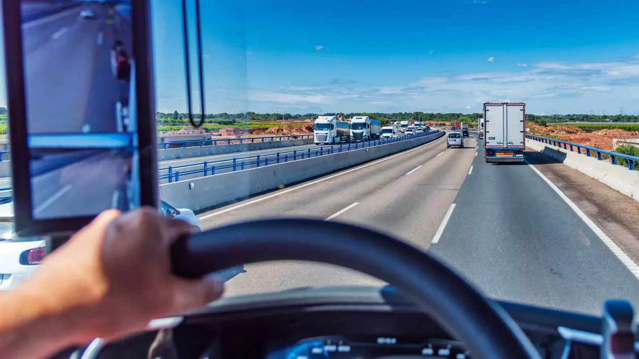 Un șofer de TIR la volanul camionului, conduce pe o autostradă într-o zi însorită, se vede că pe drum mai sunt și alte mașini.