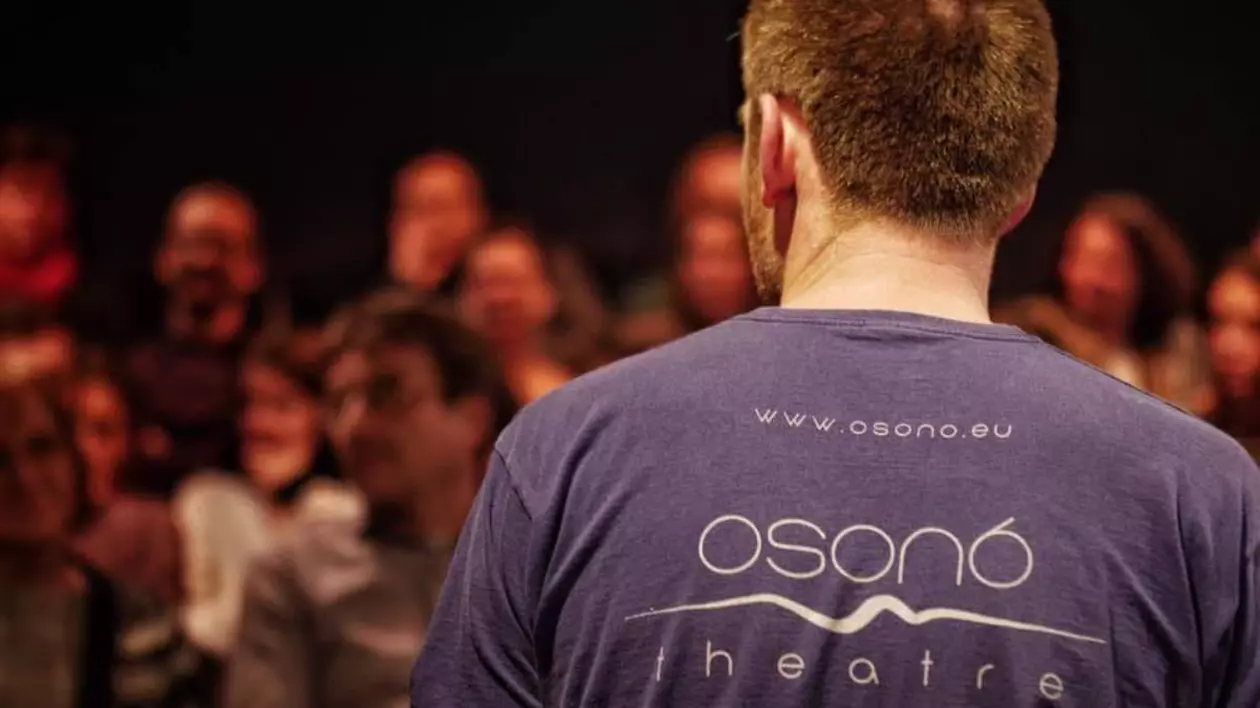 Un angajat al Teatrului Osuno în timpul unui spectacol pe scenă, în fața publicului. Bărbatul stă cu spatele la cameră și poartă un tricou pe care scrie Osuno.