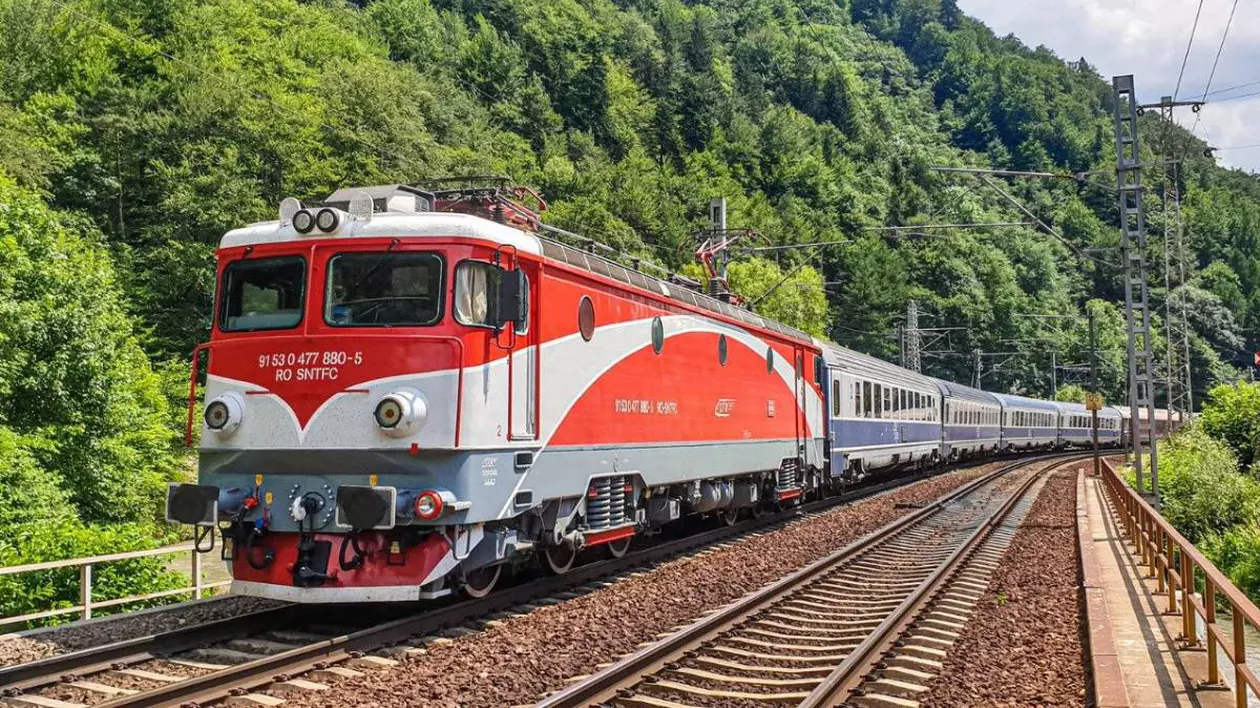 Un tren de călători cu locomotivă roșie merge pe calea ferată din România într-o zonă montană, este înconjurată linia de cale ferată de arbori cu frunze verzi.