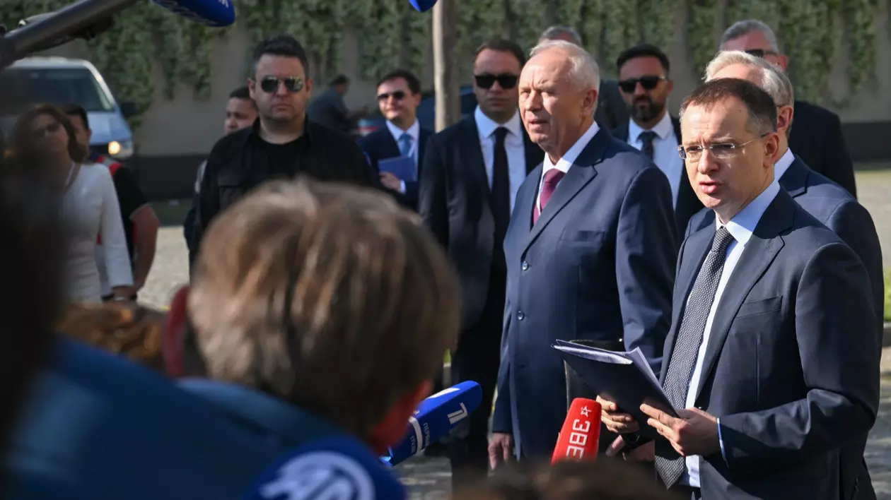 Imaginea surprinde un eveniment de presă desfășurat în aer liber, în care mai mulți oficiali, îmbrăcați în costume, dau declarații jurnaliștilor. În prim-plan, în partea dreaptă, un bărbat ține în mână un dosar cu documente și vorbește în fața mai multor microfoane