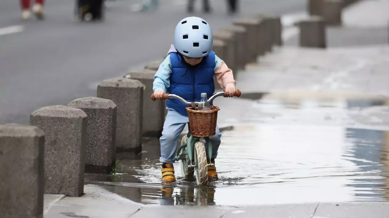 Un băiețel cu cască bleu, îmbrăcat cu mânecă lungă și vestă, este pe bicicletă și trece printr-o baltă de pe trotuar. Are adidași galbeni, iar în coșul de paie pus în fața bicicletei este o sticlă cu apă