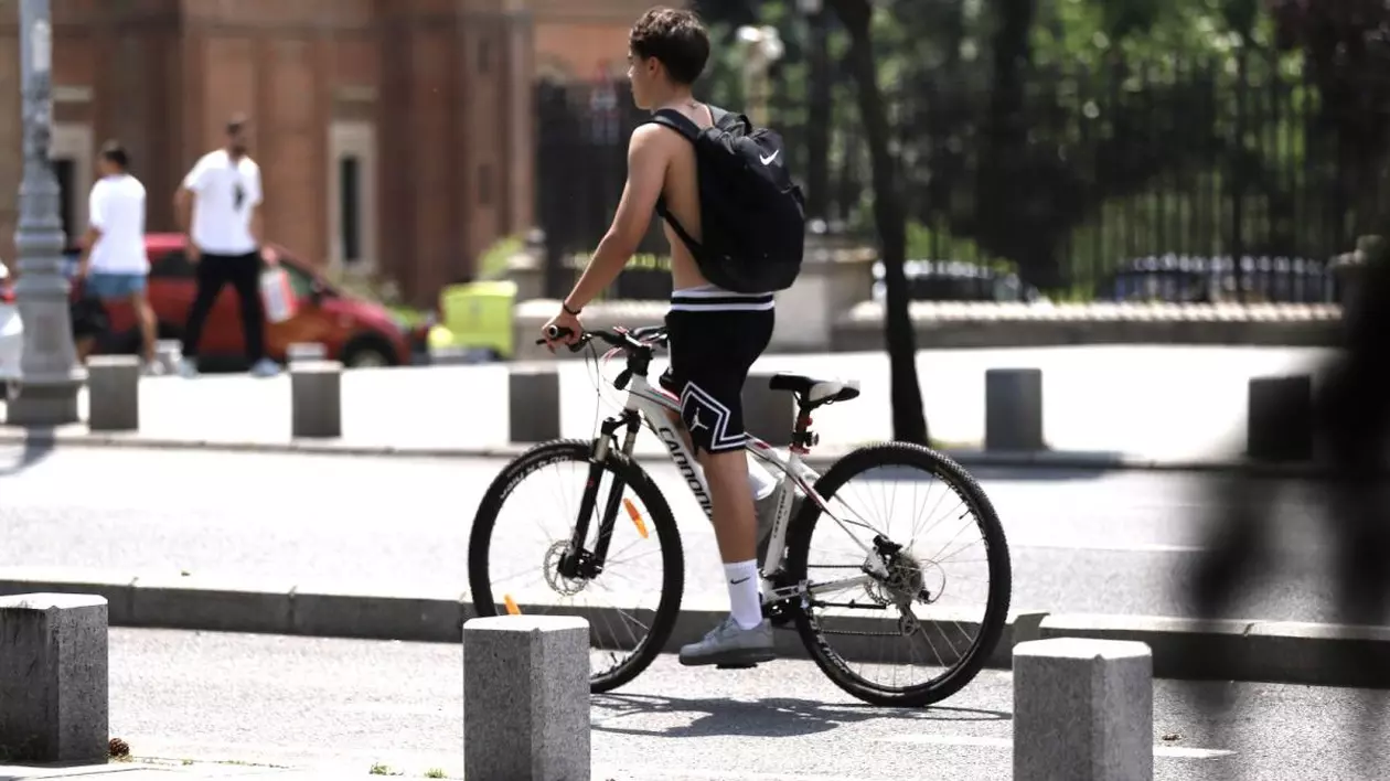 Un băiat fără tricou și cu un ghiozdan negru în spate merge pe bicicletă în picioare, într-o zi caldă de vară. În stânga se văd doi oameni în tricouri albe, iar în stânga o zonă verde dintr-un parc