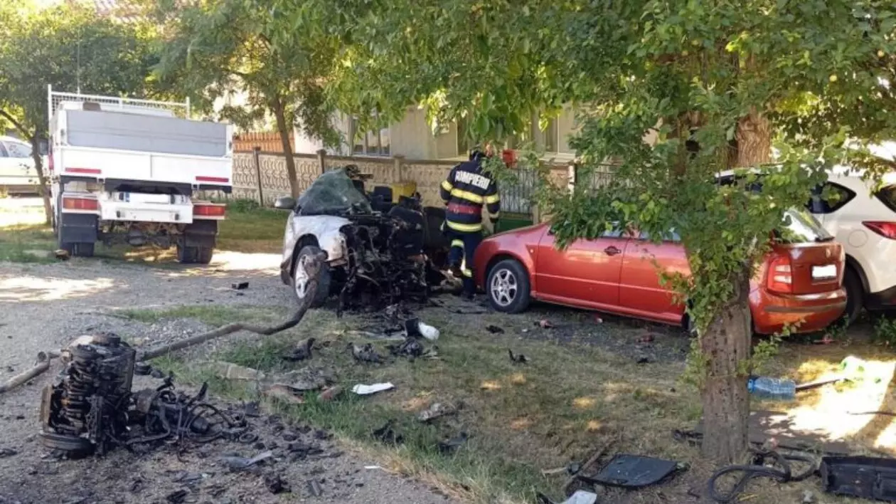un autoturism alb, complet distrus în partea din față, cu motorul și alte componente aruncate la câțiva metri distanță de caroserie. Lângă autoturism se află un pompier îmbrăcat în uniformă roșie și pe marginea drumului, sunt parcate alte două mașini, una roșie și una albă, care par să fi fost avariate ușor.