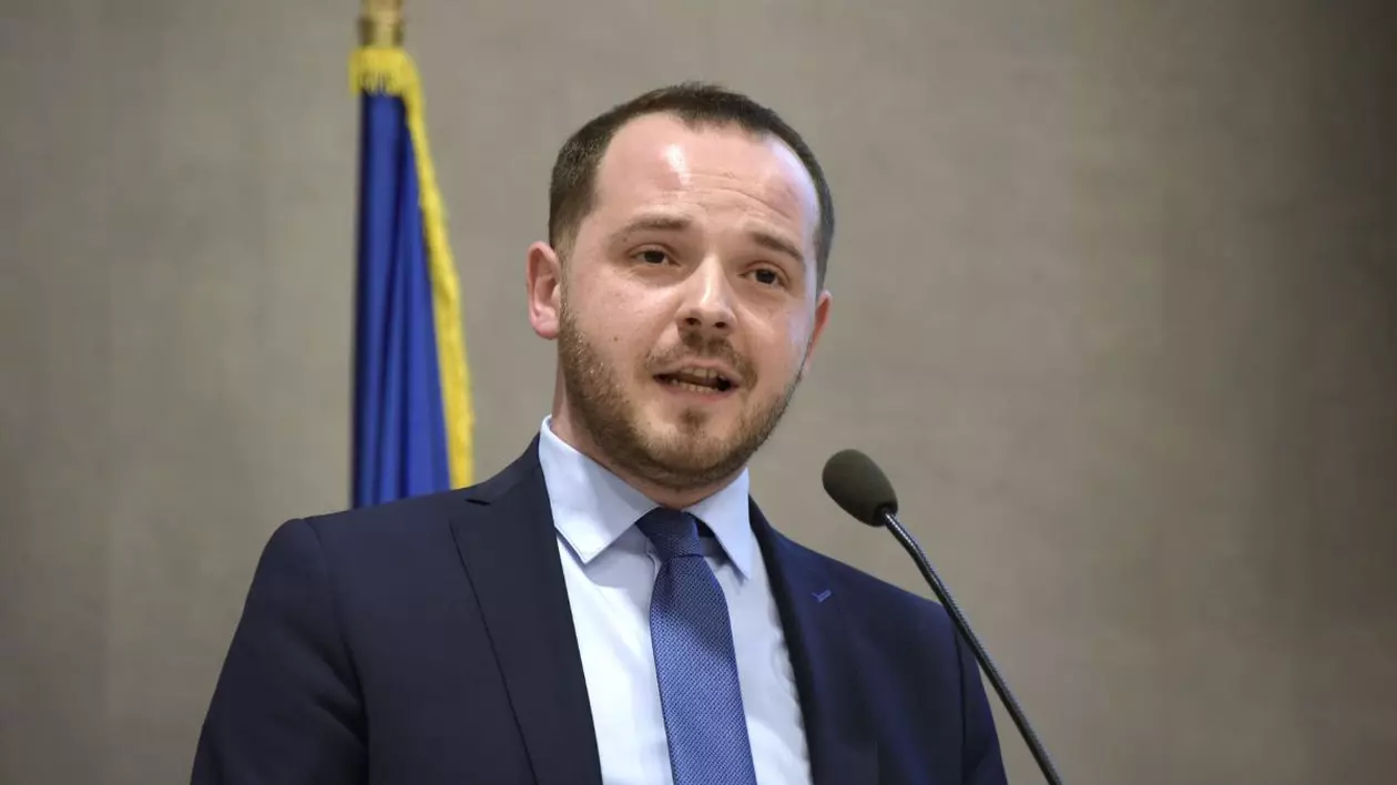 Alexandru Robodete, îmbrăcat într-un costum negru cu o cămașă albă și o cravata albastru închis, vorbind la microfon. în spatele sau se află un drapel al României