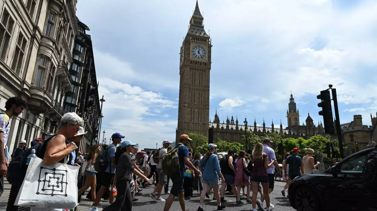 Un grup de trecători traversează strada în Londra, în fața Big Ben, într-o zi caniculată, oamenii sunt îmbrăcați subțire și se protejează de căldură cu șepci