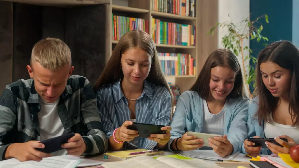 Patru elevi folosesc telefoane mobile, în timp ce sun așezați la o masă acoperită de cărți și caiete deschise, in spatele lor se vede o bibliotecă
