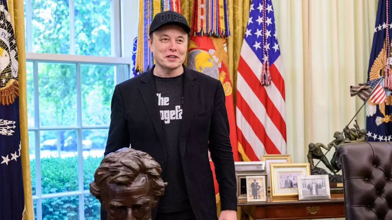 Elon Musk, stând în picioare și zâmbind, cu mâinile pe lângă corp, cu o șapcă neagră simplă pe cap, un sacou negru și un tricou negru cu textul „The Godfather” în Biroul Oval al Casei Albe, în timpul unei conferințe de presă. În soatele său se pot vedea drapelul SUA și o bibliotecă cu cărți colorate, o măsuță mică de lemn închis la culoare pe care sunt așezate mai multe rame aurii cu poze și în fața sa, în partea de stânga jos, se observă bustul de bronz al fostului președinte american Ambraham Lincoln