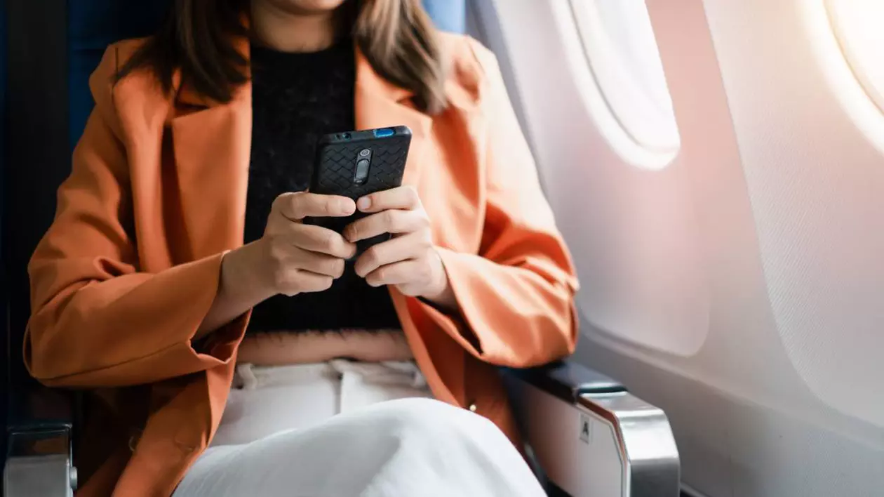 O femeie care stă cu telefonul în mână în avion și folosește internetul, poartă un palton portocaliu și pantaloni albi.