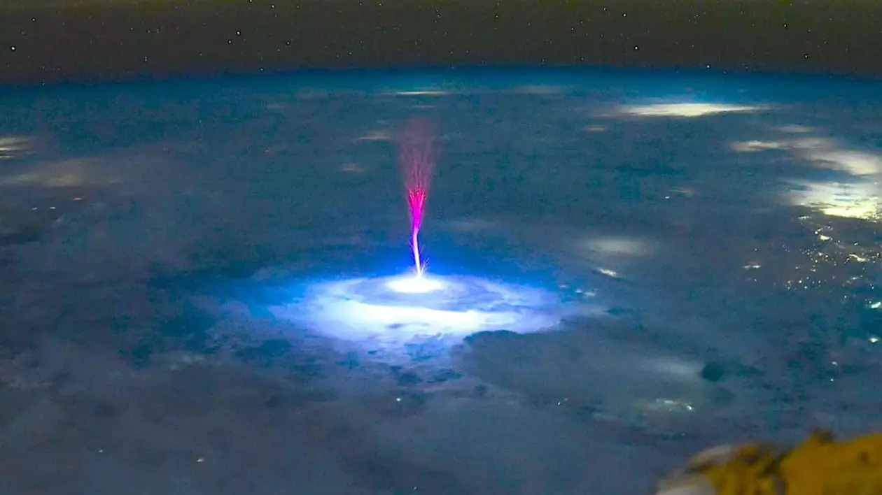 explozie de lumină albastră intensă, ca o pată circulară, care luminează norii de sub - în mijlocul acestei zone luminoase pornește un jet vertical de lumină roșie-violet, care se ridică în sus spre cer